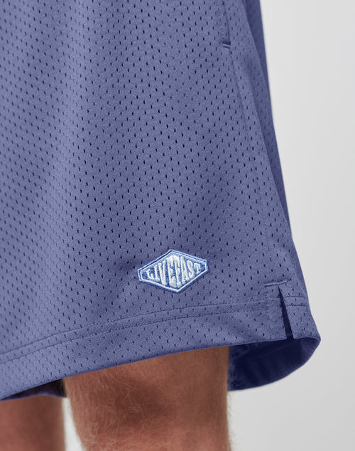 Close-up of a person wearing blue Basic Mesh Shorts by LIVE FAST DIE YOUNG, featuring an elastic waistband and a small logo patch near the hem. Focus is on the mesh texture, regular fit, and brand logo on the lower left side.