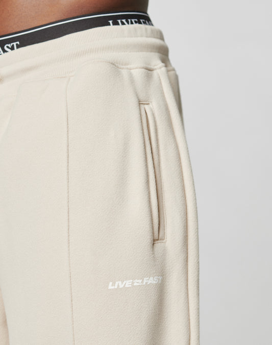 Close-up of a person wearing beige LFDY Pleated Jogger by LIVE FAST DIE YOUNG, featuring a vertical pocket and white LIVE FAST logo. The black elastic waistband has white LIVE FAST text. Only the lower torso and upper legs are shown.