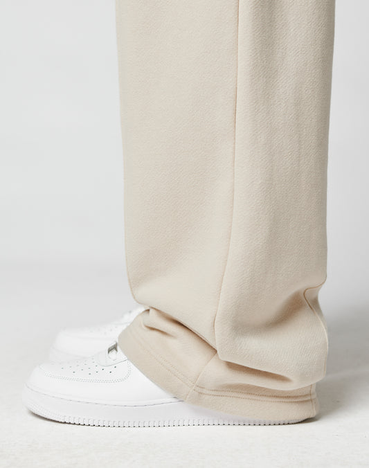 A close-up shows a person wearing LIVE FAST DIE YOUNGs LFDY Pleated Jogger in wide fit beige, featuring an Elastik-Bund, draped over clean white sneakers against a simple white background.