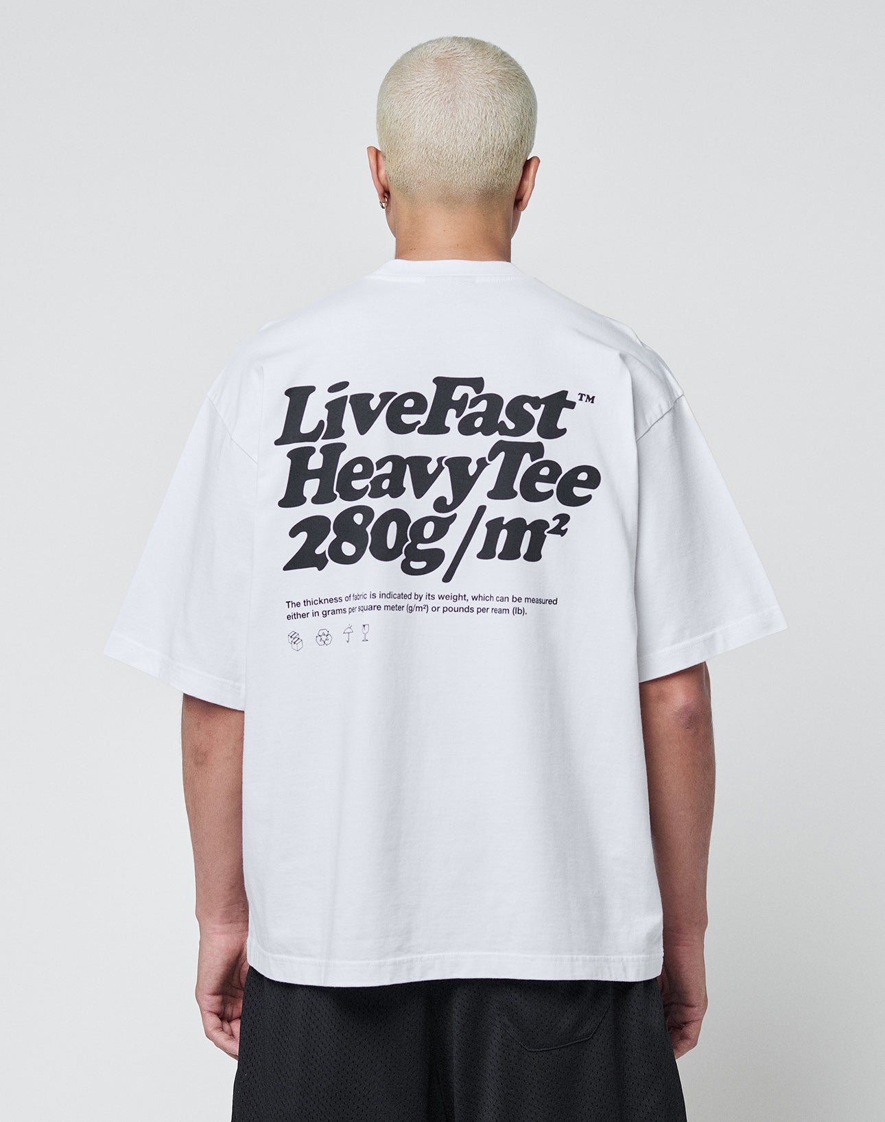 A person with short, bleached hair is seen from behind wearing a white LIVE FAST DIE YOUNG Basic 280 Tee with bold black text and symbols, paired with black shorts.