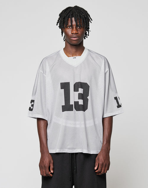 A man with short dreadlocks wears the LIVE FAST DIE YOUNG LFDY Mesh Jersey—an oversized, light gray polyester jersey with a V-neck and black number 13 on the front and sleeves—against a plain light background.
