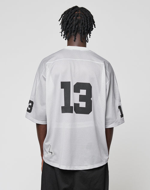 A person with short twisted hair stands with their back to the camera, wearing the oversize-fit white LFDY Mesh Jersey by LIVE FAST DIE YOUNG featuring a V-neck and black number 13, paired with black shorts.