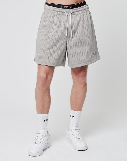 A person stands against a plain white background wearing LIVE FAST DIE YOUNG Basic Mesh Shorts (gray, logo patch, regular fit), white LIVE FAST socks, white sneakers, and black LIVE FAST underwear. Upper body not visible.