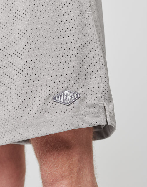 Close-up of a persons leg wearing LIVE FAST DIE YOUNG Basic Mesh Shorts in light gray, 100% polyester, regular fit, with a small diamond-shaped logo patch near the hem.