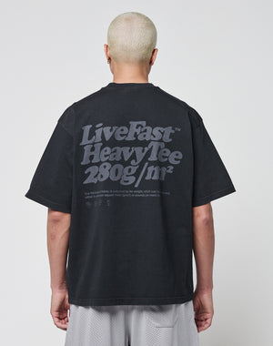 A person with short, bleached blonde hair is shown from behind, wearing a black LIVE FAST DIE YOUNG Basic 280 Tee with a bold backprint. The background is plain white.