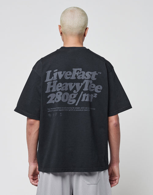 A person with short, bleached blonde hair is shown from behind, wearing a black LIVE FAST DIE YOUNG Basic 280 Tee with a bold backprint. The background is plain white.