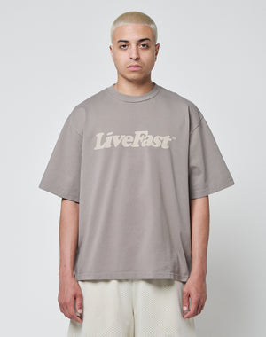 A person with short, bleached blonde hair wears the LIVE FAST DIE YOUNG Basic Tee in taupe, Oversize Fit aus Baumwolle mit Vintage-Waschung and LiveFast print, paired with light mesh shorts, against a plain white background.