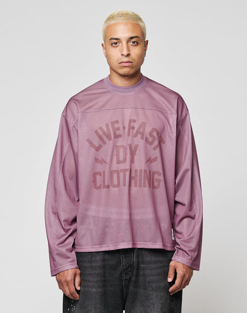 A person with short blond hair wears the LFDY Flash Jersey by LIVE FAST DIE YOUNG—an oversize, long-sleeve mauve mesh top with a bold LIVE FAST DIY CLOTHING print—paired with dark jeans, standing against a plain light gray background.