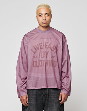 A person with short blond hair wears the LFDY Flash Jersey by LIVE FAST DIE YOUNG—an oversize, long-sleeve mauve mesh top with a bold LIVE FAST DIY CLOTHING print—paired with dark jeans, standing against a plain light gray background.