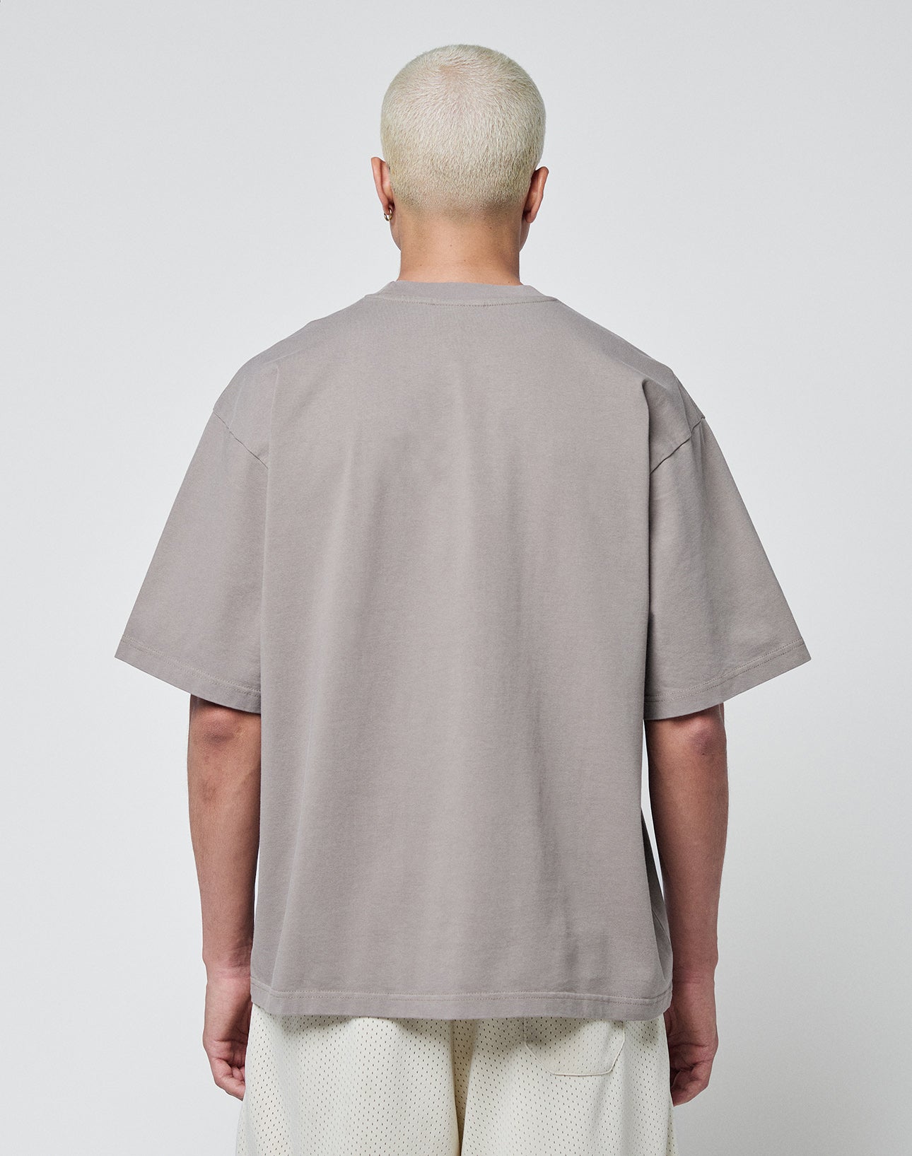 A person with short, bleached blond hair is standing with their back to the camera, wearing a LIVE FAST DIE YOUNG Basic Tee and white shorts against a plain light background.