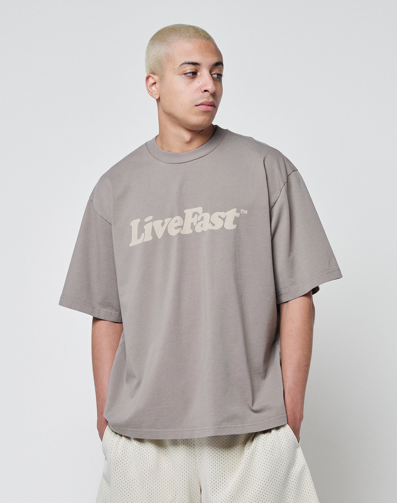 A person with short blond hair wears the LIVE FAST DIE YOUNG Basic Tee in Oversize Fit, featuring LiveFast on the front, made from soft cotton, paired with light mesh shorts, standing against a plain white background.
