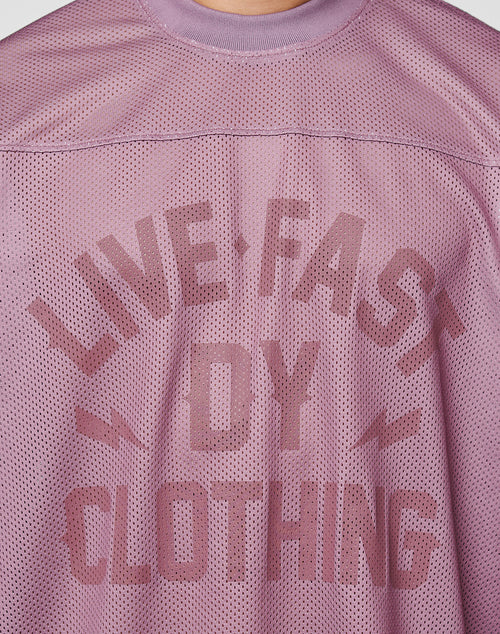 A close-up of the LFDY Flash Jersey by LIVE FAST DIE YOUNG shows its mauve mesh fabric, bold LIVE FAST BY CLOTHING text, lightning bolt graphics, and oversize fit for a modern streetwear vibe.