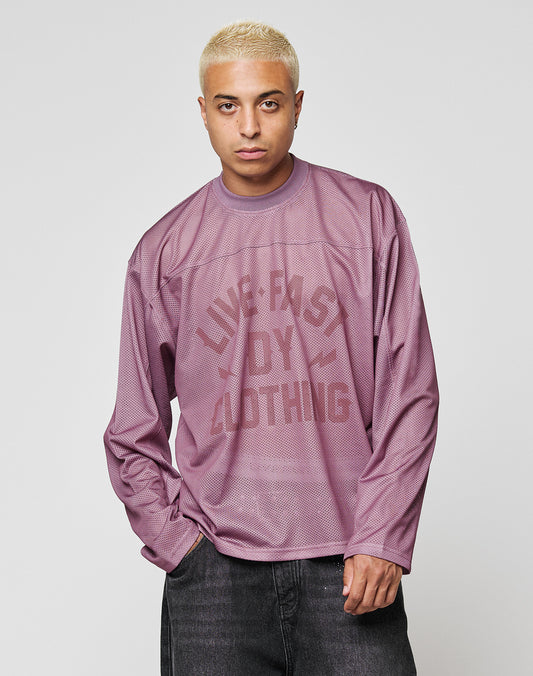A short-haired blond person wears the LFDY Flash Jersey by LIVE FAST DIE YOUNG—an oversized mauve mesh top with a bold front print—standing with one hand in their pocket against a plain light gray background.