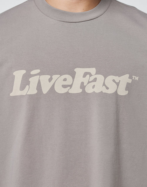 A close-up of a person wearing the LIVE FAST DIE YOUNG Basic Tee, a gray T-shirt with vintage wash and LiveFast™ printed in bold, beige letters across the chest.