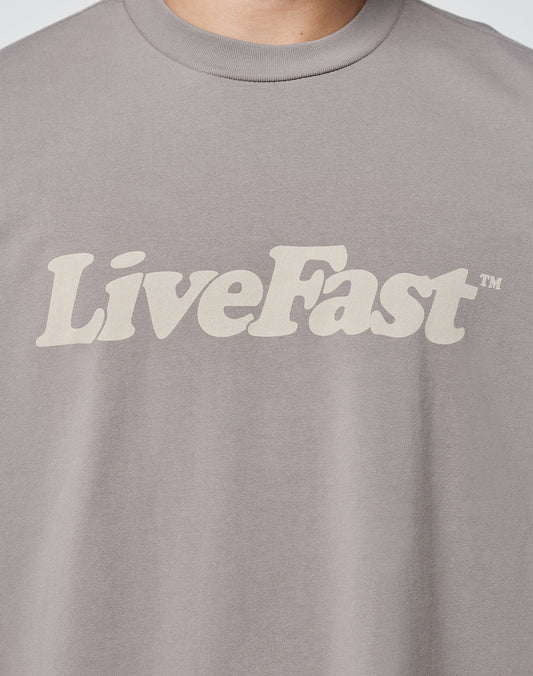 A close-up of a person wearing the LIVE FAST DIE YOUNG Basic Tee, a gray T-shirt with vintage wash and LiveFast™ printed in bold, beige letters across the chest.