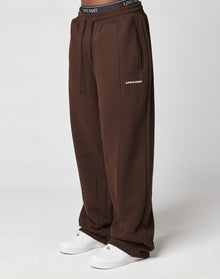 Someone wearing LIVE FAST DIE YOUNG LFDY Pleated Jogger wide fit brown sweatpants with LIVE FAST text on the left thigh, elastic waistband, and white sneakers, stands against a plain light background.