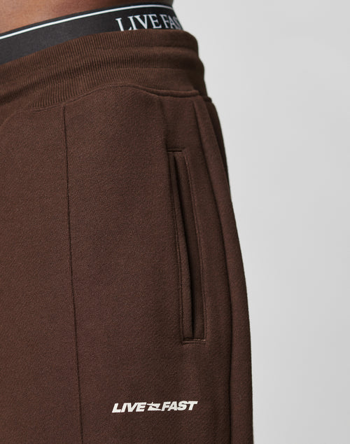 Close-up of a person wearing dark brown LFDY Pleated Jogger by LIVE FAST DIE YOUNG, featuring a side pocket and white LIVE FAST print near the pocket and on the elastic waistband. Plain light-colored background.