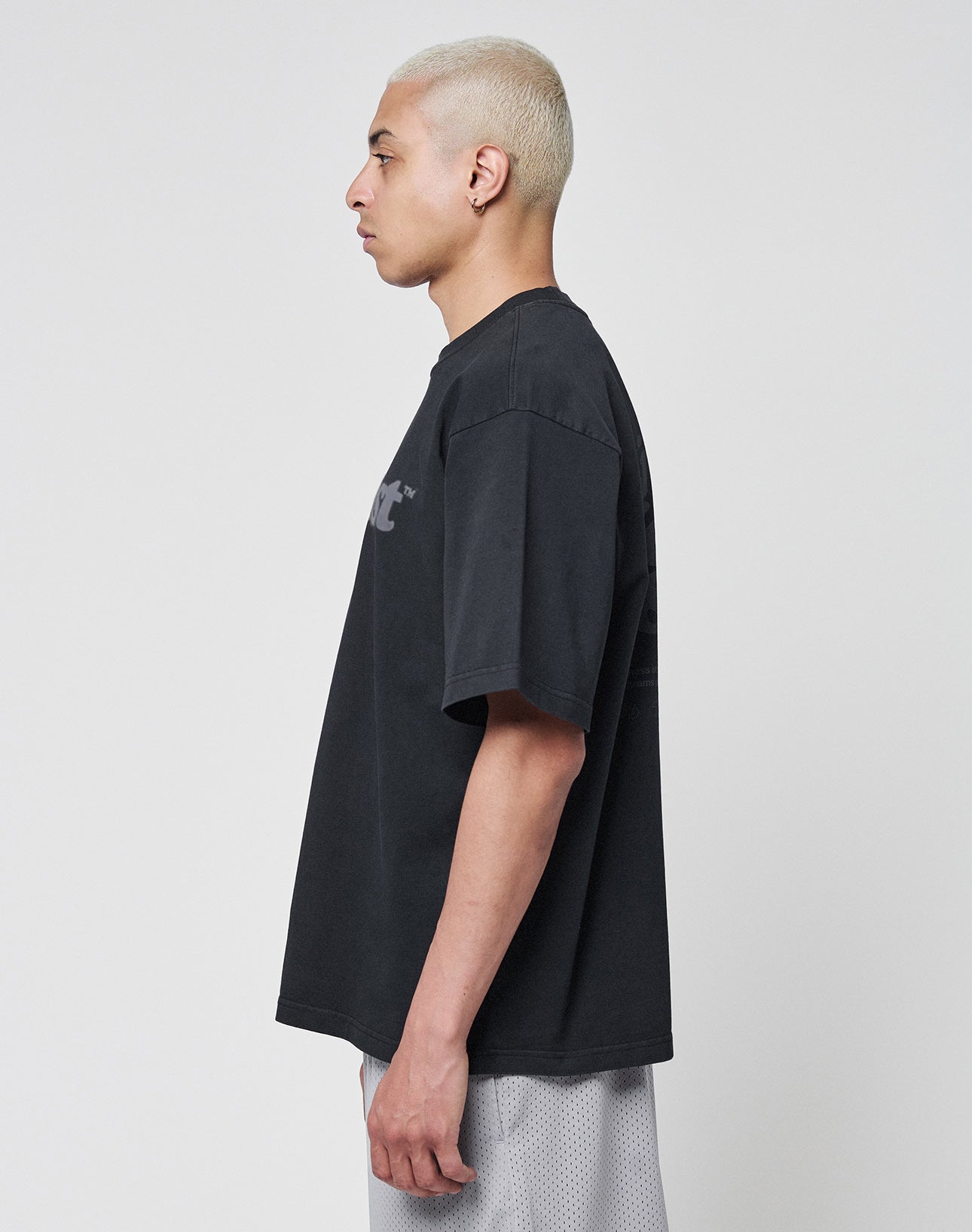 A person with short bleached blond hair stands in profile against a plain white background, wearing the LIVE FAST DIE YOUNG Basic Tee aus 100% Baumwolle and light-colored shorts.