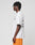 A person with short braided hair stands in profile wearing the LIVE FAST DIE YOUNG LFDY Praying Tee—an oversize white graphic t-shirt with front and back print—and orange athletic shorts against a plain light background.