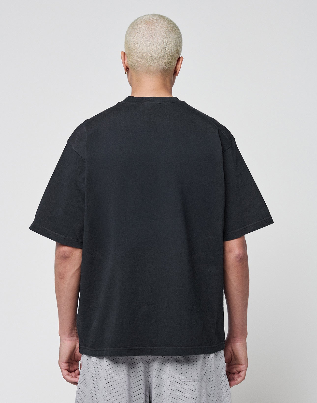 A person with short, bleached blond hair stands with their back to the camera, wearing a LIVE FAST DIE YOUNG Basic Tee in Oversize Fit and light gray mesh shorts against a plain light background.