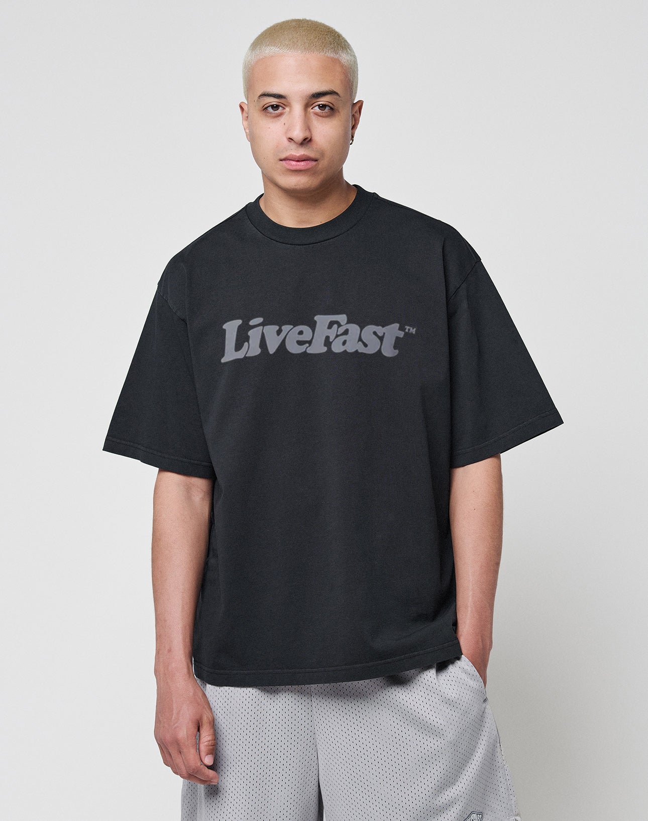 A person with short, bleached blond hair wears a LIVE FAST DIE YOUNG Basic Tee in an oversize fit with bold LiveFast lettering, paired with light gray mesh shorts, against a plain white background.