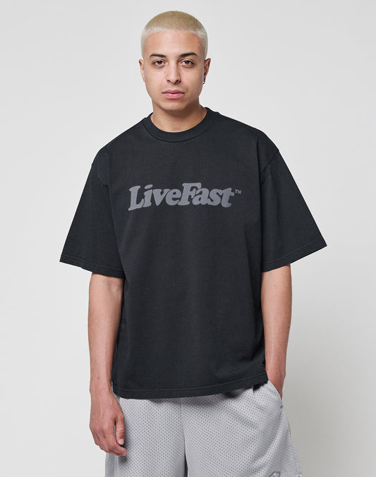 A person with short, bleached blond hair wears a LIVE FAST DIE YOUNG Basic Tee in an oversize fit with bold LiveFast lettering, paired with light gray mesh shorts, against a plain white background.