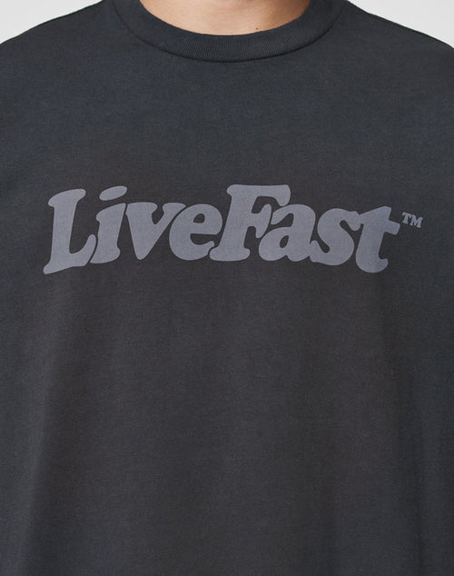 Someone wears the Basic Tee by LIVE FAST DIE YOUNG—a black T-shirt with bold gray LiveFast™ text, featuring an oversize fit and vintage wash. Made from 100% Baumwolle for all-day comfort.
