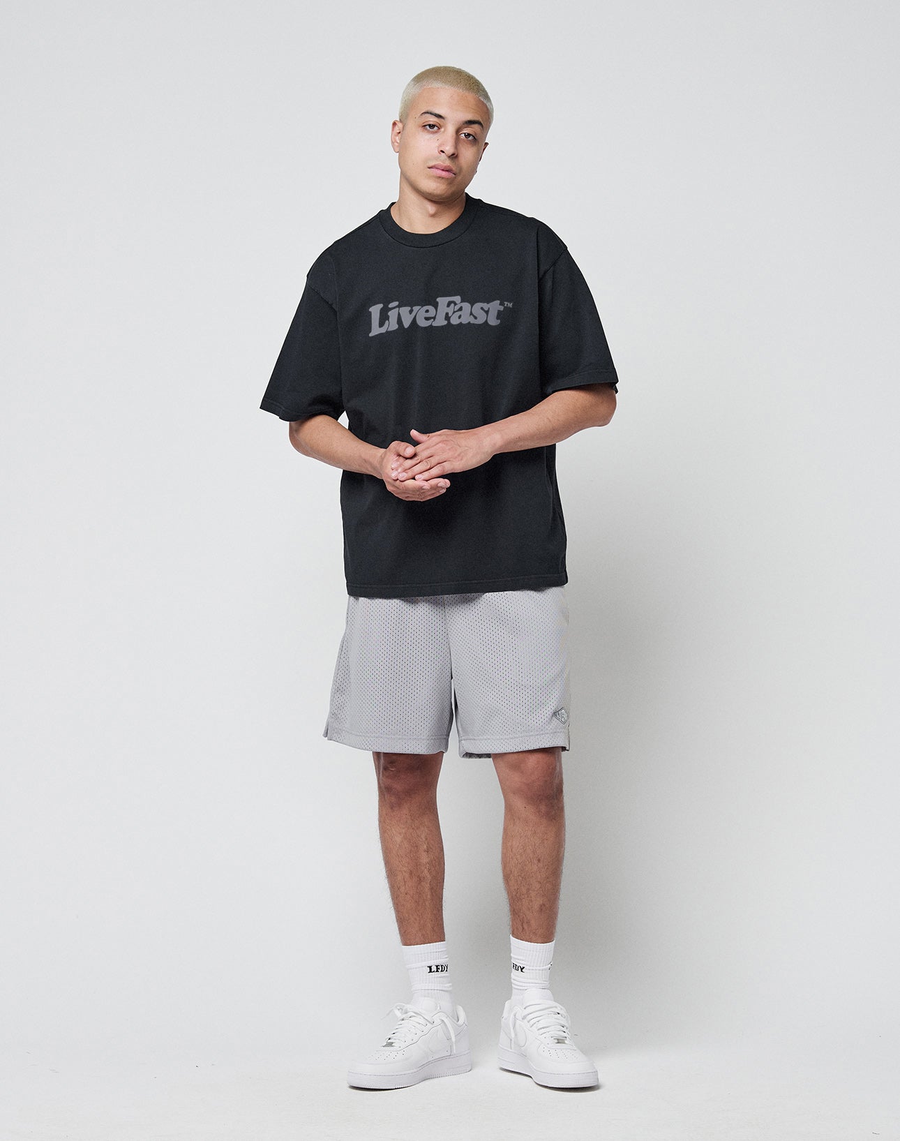 A person with short blond hair stands against a plain background, wearing the LIVE FAST DIE YOUNG Basic Tee in black (100% cotton, oversize fit) and light gray shorts. They have a relaxed expression, white socks, sneakers, and clasped hands.