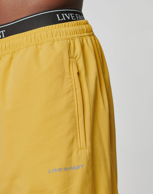 Close-up of a person wearing LFDY Trackpants by LIVE FAST DIE YOUNG with yellow polyester fabric, Elastikbund, and LIVE FAST as reflektierender Druck near the pocket and waistband. Plain light background highlights the pants.