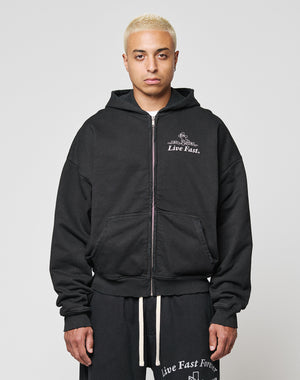 A person with short bleached blonde hair wears the LFDY Praying Hooded Zip by LIVE FAST DIE YOUNG, styled with black sweatpants featuring white drawstrings and Live Fast and Live Fast Forever text. The background is plain gray.