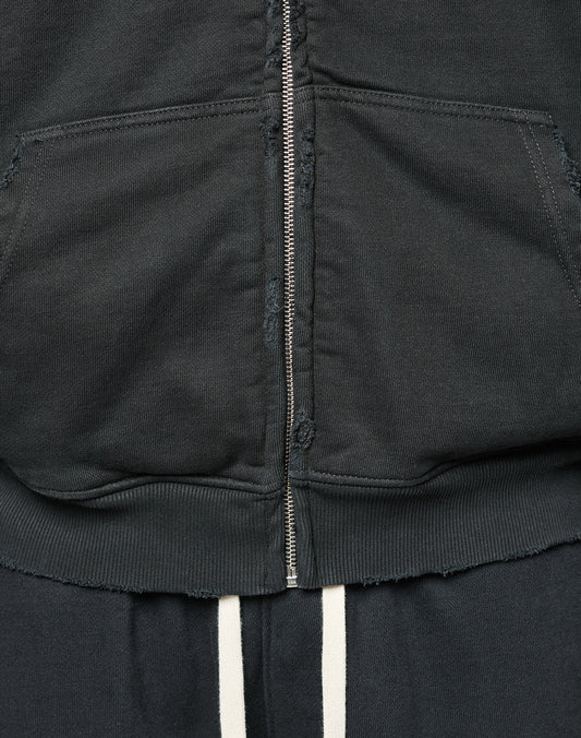 Close-up of the LFDY Praying Hooded Zip by LIVE FAST DIE YOUNG, a black oversized hoodie with a metal zipper, front pouch pocket, frayed hem Distress-Effekt, paired with dark pants featuring two vertical white stripes.