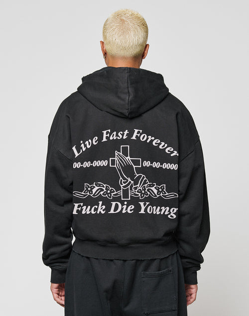 A person with short, bleached hair wears the LIVE FAST DIE YOUNG LFDY Praying Hooded Zip, an oversized hoodie with a distressed look, Live Fast Forever, Fuck Die Young text on the back, plus praying hands and cross graphics.