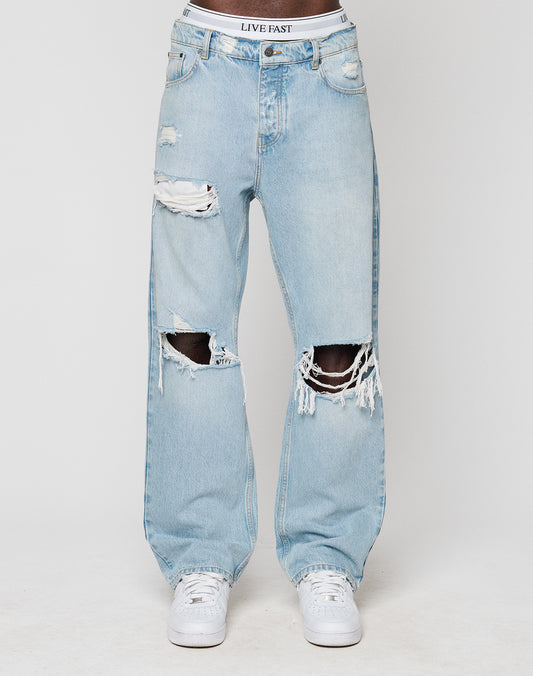 A person wears LIVE FAST DIE YOUNGs LFDY Distressed Baggy Denim—light blue, high-waisted, 100% Baumwolle jeans with large rips and frayed edges—paired with white sneakers and visible LIVE FAST branded underwear.