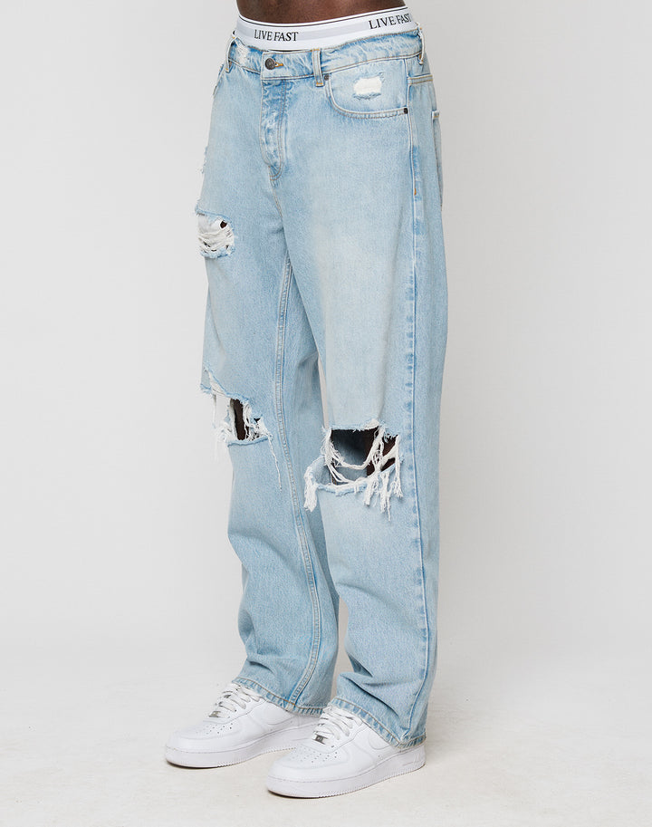 A person stands against a plain white background wearing LIVE FAST DIE YOUNG’s LFDY Distressed Baggy Denim—light blue jeans with heavy distressing, large rips on both legs, white sneakers, and a waistband reading LIVE FAST.