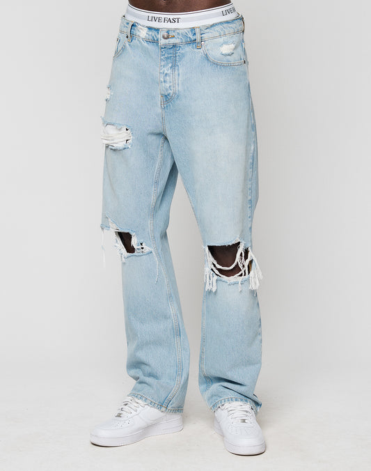 Someone wears LFDY Distressed Baggy Denim from LIVE FAST DIE YOUNG—light blue, 100% Baumwolle jeans with large knee rips, frayed edges, a baggy fit, exposing skin and LIVE FAST waistband, styled with white sneakers.