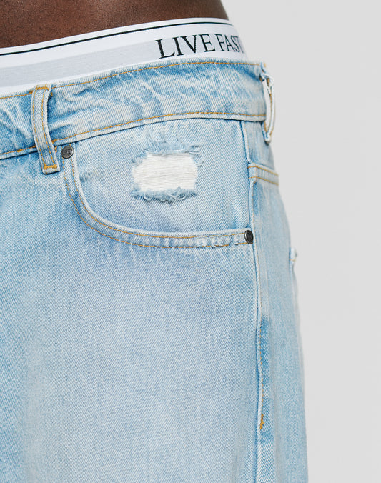 Close-up of the LFDY Distressed Baggy Denim by LIVE FAST DIE YOUNG. Light blue, 100% Baumwolle jeans with a ripped detail near the pocket and a baggy fit showing white underwear printed with LIVE FAST.
