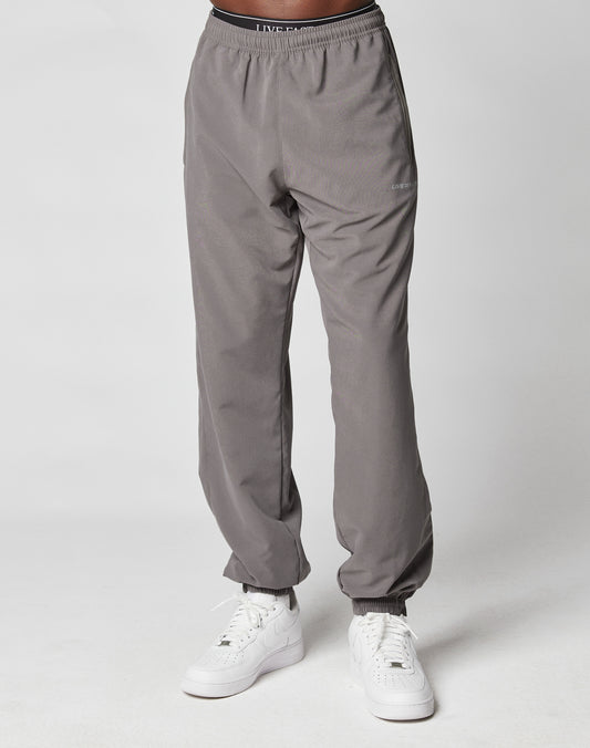 A person wears LIVE FAST DIE YOUNG LFDY Trackpants with Elastikbund and white sneakers, standing against a plain white background. Only the lower half is visible, highlighting the pants casual Regular Fit.