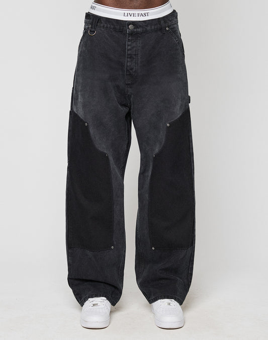 A person showcases the Carpenter Look in loose-fitting black LFDY Carpenter Pants by LIVE FAST DIE YOUNG, with large dark thigh patches, white sneakers, and a visible LIVE EAST waistband against a plain light background.