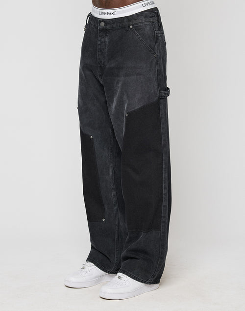A person models the classic Carpenter Look in baggy-fit, black LFDY Carpenter Pants by LIVE FAST DIE YOUNG, featuring large dark knee patches and a LIVE FAST waistband, paired with white sneakers. The background is plain and light-colored.