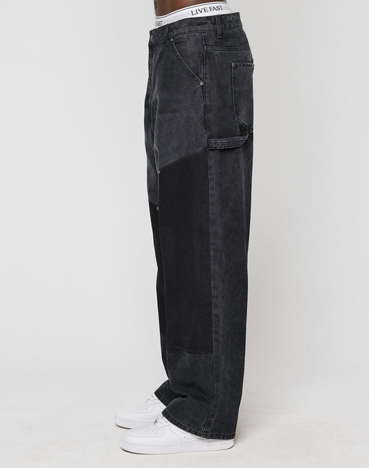 Model wears LFDY Carpenter Pants by LIVE FAST DIE YOUNG—baggy fit black denim with carpenter style, side pocket, and white waistband showing. Paired with white sneakers, standing sideways against a plain background. Made from 100% Baumwolle.
