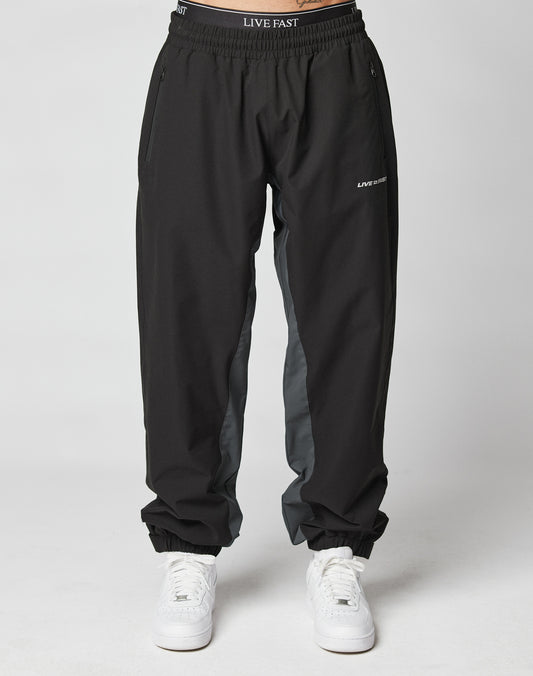 A person wears LIVE FAST DIE YOUNG Basic Shell Pants—black joggers with gray side panels, elastic drawstring waist reading “LIVE FAST,” zippered side pockets, and a reflective logo on the left thigh. Styled with white sneakers; plain white background.