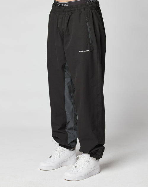 A person wears LIVE FAST DIE YOUNG Basic Shell Pants with an elastic waistband and drawstring, “LIVE FAST” reflective print near the left pocket, a gray inner leg stripe, and white sneakers, standing against a plain light background.