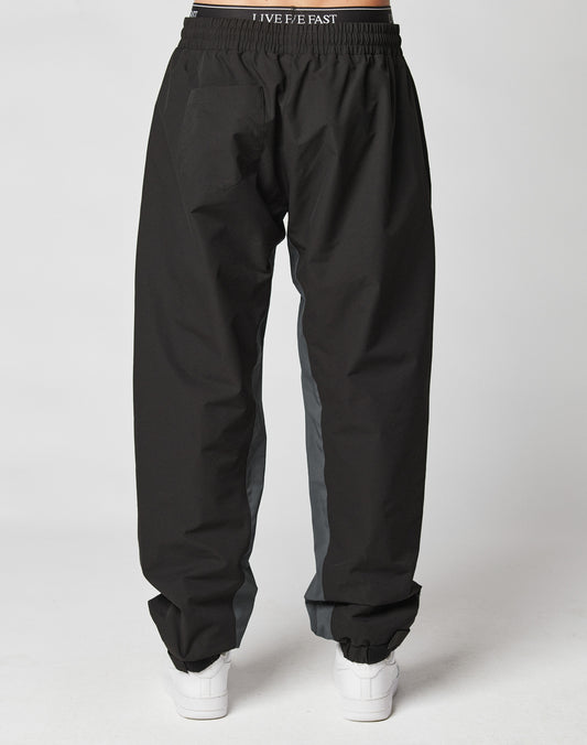 A person wearing LIVE FAST DIE YOUNG Basic Shell Pants, loose black with gray side panels and an elastic drawstring waistband, styled with white sneakers, photographed from the back against a plain white background.