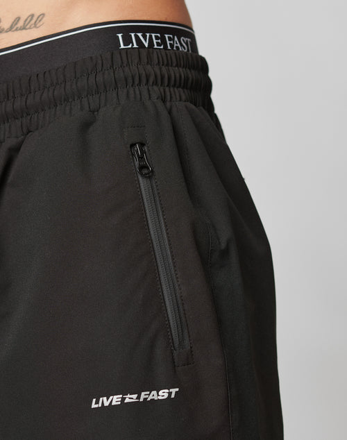 Close-up of a person wearing LIVE FAST DIE YOUNG Basic Shell Pants in black with an oversize fit, zippered pocket, and reflective LIVE FAST print on the waistband and leg. Lower abdomen and waist are also visible.