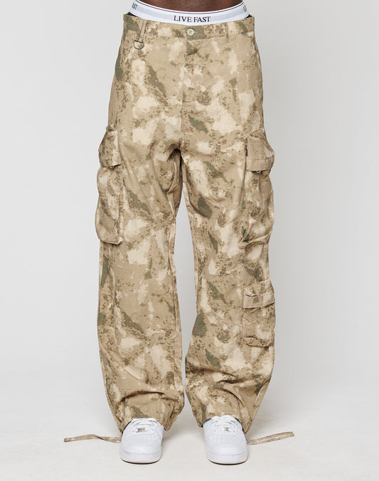 A person wears LFDY Cargo pants from LIVE FAST DIE YOUNG in wide fit tan camo with multiple cargo pockets and a visible LIVE FAST waistband. Made from 100% cotton, styled with white sneakers against a plain white background.