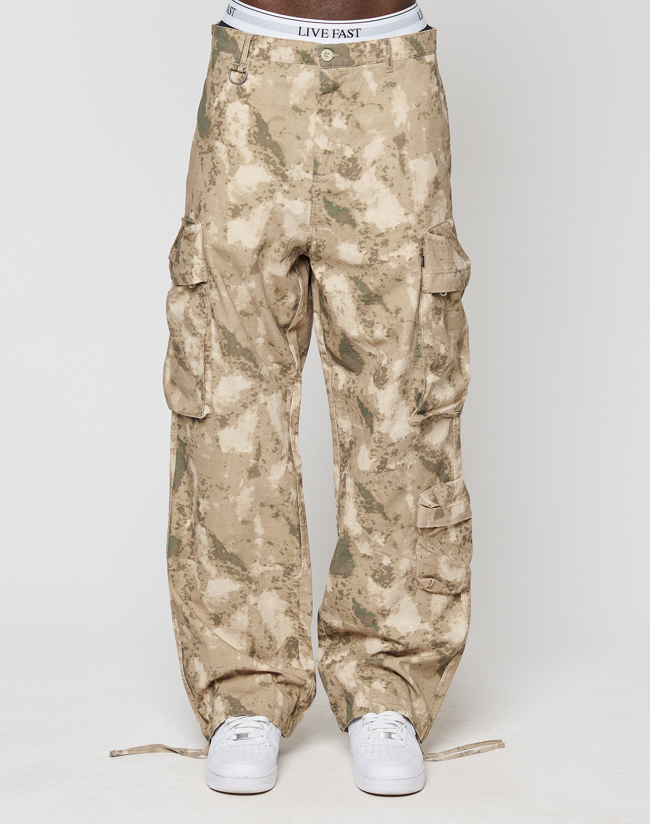 A person in LFDY Cargo pants by LIVE FAST DIE YOUNG—wide fit, tan camo print, 100% cotton, with cargo and side pockets—white LIVE FAST waistband visible, paired with white sneakers, against a plain white background.