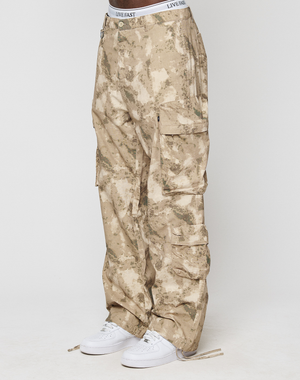 A person wears beige and tan camouflage LFDY Cargo pants from LIVE FAST DIE YOUNG, paired with white sneakers and a white LIVE FAST waistband, standing against a plain white background.