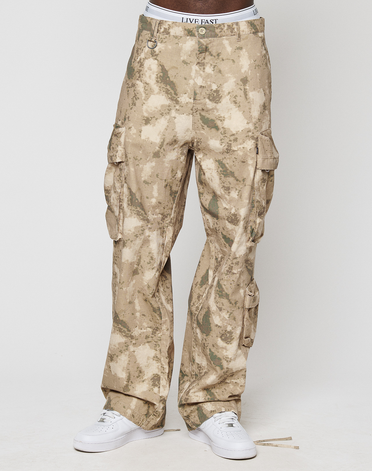 A person is wearing beige camo LFDY Cargo pants by LIVE FAST DIE YOUNG with multiple cargo pockets and "LIVE FAST" on their underwear waistband, paired with white sneakers, standing against a plain white background. Upper body not visible.