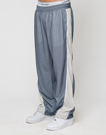 A person wears blue-gray LFDY DY Trackpants by LIVE FAST DIE YOUNG with a wide fit, elastic waistband reading "LIVE FAST," white side stripes, and white sneakers, standing against a plain light background.