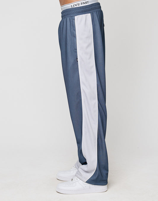 Side view of a person in LFDY DY Trackpants by LIVE FAST DIE YOUNG—wide fit blue polyester with white side stripe, elastic waistband featuring LIVE FAST text, paired with white sneakers against a plain background.
